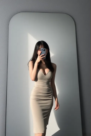 거울 앞에서 베이지색 슬림 원피스를 착용한 여성이 휴대전화로 셀피를 찍는 모습